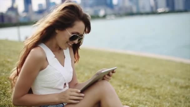 Ein attraktives Mädchen mit langen braunen Haaren, weißem Hemd, Jeanshose und schwarzer Sonnenbrille sitzt im Gras an einem See und hält ein Tablet in der Hand — Stockvideo
