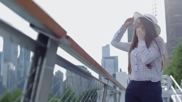 Una mujer joven con sombrero y gafas de sol está parada bajo el sol en el paseo marítimo y hablando por teléfono . — Vídeos de Stock