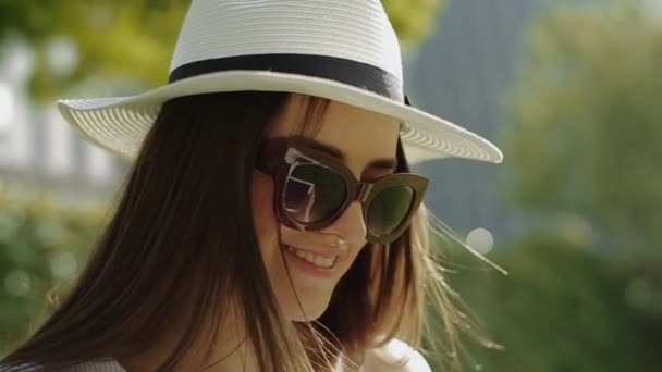 Junge schöne lächelnde Frau mit Hut und Sonnenbrille, glättet ihre Haare und sieht direkt im Park in der Mitte aus. — Stockvideo