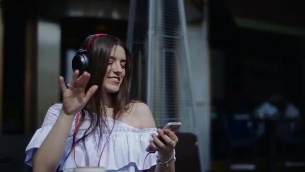 Sorrindo jovem mulher com fones de ouvido apreciando música dançando em cafés de rua . — Vídeo de Stock