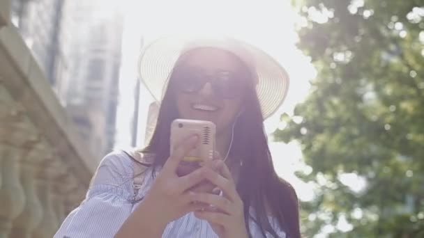 Mujer joven con sombrero y gafas de sol hace una videollamada desde su teléfono inteligente en el centro de la ciudad . — Vídeos de Stock