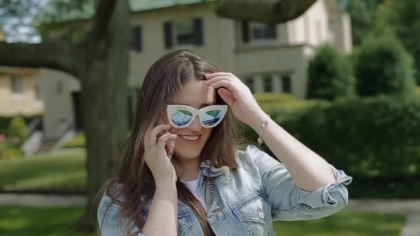 Jonge aantrekkelijke vrouw in zonnebril op telefoon praten en rechtzetten van lange haren in een woonwijk met twee verdiepingen huizen. — Stockvideo