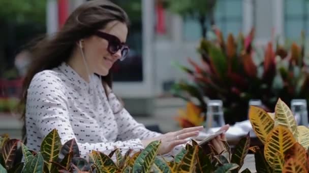 Mladá krásná žena v sluneční brýle sedí na chodníku Cafe mezi okrasné rostliny. — Stock video