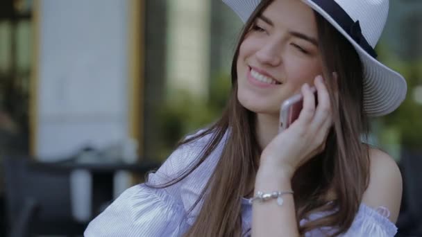 Joven, sonriente mujer de sombrero, está llamando por teléfono sentado en una mesa en un café al aire libre . — Vídeos de Stock