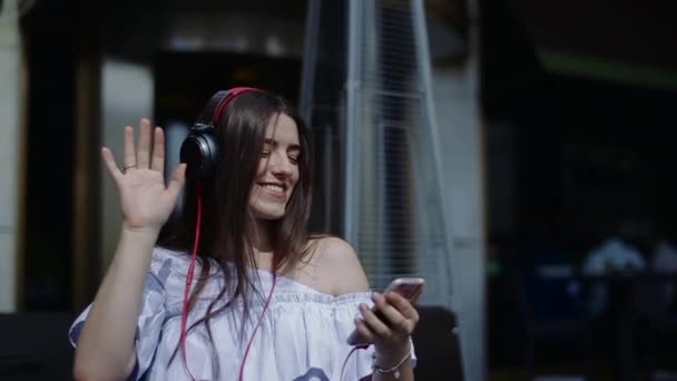 Sorrindo jovem mulher com fones de ouvido apreciando música dançando em cafés de rua . — Vídeo de Stock