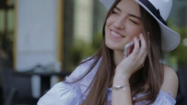 年轻、 微笑的女人，戴着帽子，呼吁手机坐在一张桌子上户外咖啡厅. — 图库视频影像