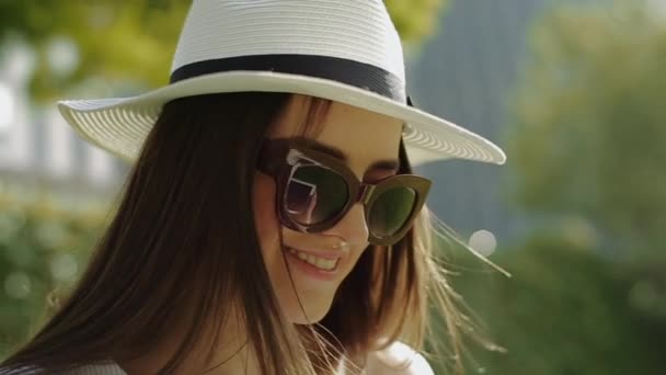 Joven hermosa mujer sonriente en sombrero y gafas de sol, endereza su cabello y se ve justo en el parque en el centro . — Vídeo de stock