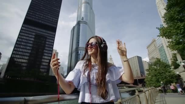 Junge schöne Frau hört über Kopfhörer zu und tanzt auf einem Damm mit Wolkenkratzern und einem Kanal. — Stockvideo