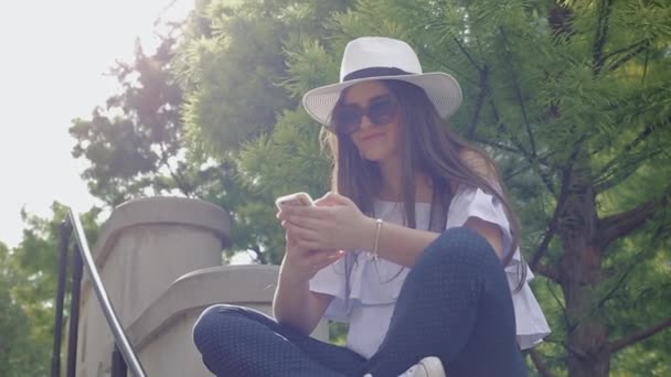 Joven hermosa mujer en sombrero en el teléfono, en el centro de la ciudad, sentado en barandilla en el parque bajo un árbol . — Vídeos de Stock
