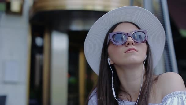 Junge schöne Frau mit Hut und Brille, die Wolkenkratzer im Gespräch auf Headset reflektieren. — Stockvideo