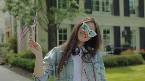 Wanita muda berkacamata melambaikan bintang dan garis-garis bendera di depan rumah dua tingkat di lingkungan . — Stok Video