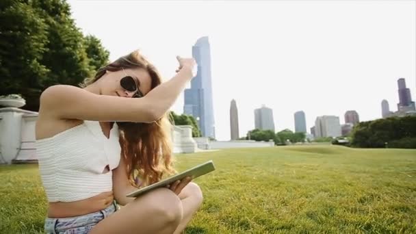 Una ragazza con lunghi capelli castani è seduta sull'erba in un parco a giocare con la sua tavoletta in una giornata soleggiata — Video Stock