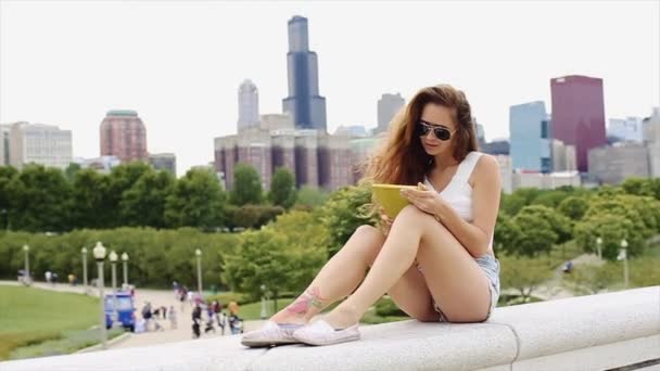 Piękna dziewczyna z białą koszulkę i szorty Jean jest siedzi i gra z urządzenia w jej ręce z parku i Downtown w tle Slow Motion — Wideo stockowe