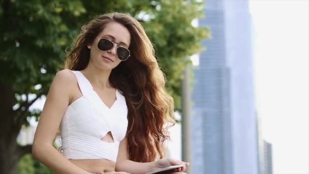 Uzun boylu binaları ile arka planda Tablet siyah güneş gözlüğü ve uzun saç oturan güzel kızla tezgah ve onunla oynamak — Stok video
