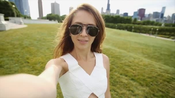 Una ragazza in un parco con lunghi capelli castani e occhiali da sole neri sta prendendo un selfie in un giorno luminoso e soleggiato — Video Stock