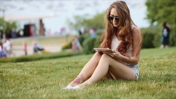 Linda chica de camisa blanca y pantalones cortos de Jean en sentarse en la hierba con su dispositivo y la gente, el lago y los edificios altos en el fondo — Vídeos de Stock