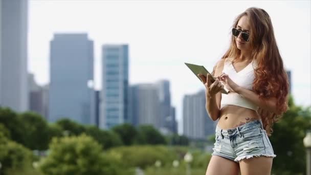 Pretty Caucasian Girl en camisa blanca y pantalones cortos Jean está jugando con el dispositivo con edificios altos, lago y árboles en el fondo en cámara lenta — Vídeo de stock