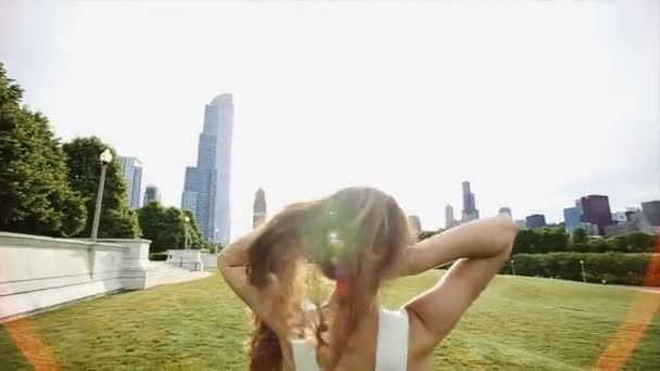 女孩与长棕色头发穿着一件白衬衫、 牛仔短裤和黑色太阳镜正在运行且在公园的慢动作跳舞 — 图库视频影像
