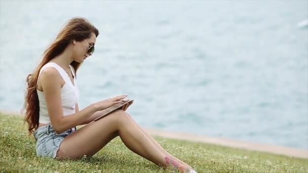 Schattige Brunette meisje zit door de Lake met behulp van haar Tablet en Her Hair bewegen rond op een zonnige dag — Stockvideo