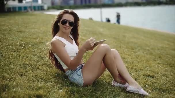 有吸引力的女孩与长棕色头发穿一件白衬衫和牛仔短裤和黑色太阳镜抱着平板电脑坐在湖边草地上 — 图库视频影像