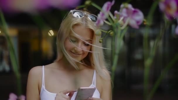 Krásná mladá dívka s brýlemi na hlavě květ orchideje se dívá na její mobilní telefon a usmívá se. Nastaví telefon tak, aby se fotografie. — Stock video