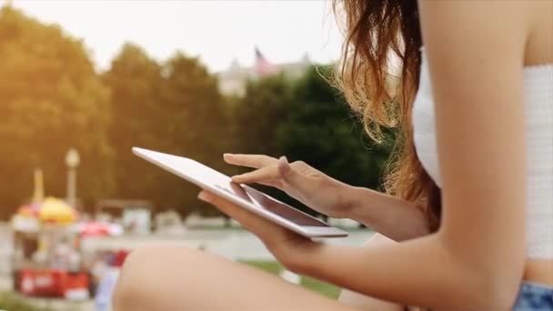 Söt flicka i vit topp och Jean Shorts med långt brunt hår är sitter och leker med hennes enhet med höga Buiding i bakgrunden Slow Motion — Stockvideo