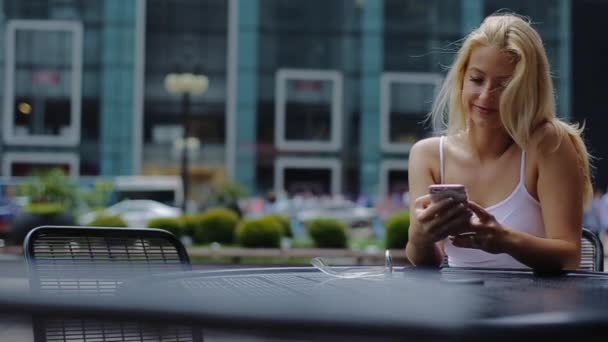 Młody atrakcyjny Blond dziewczyna siedzi przy stole w Cafe, trzymając telefon komórkowy i tarcze wiadomości. — Wideo stockowe