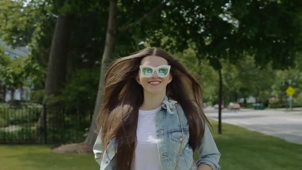 Mujer joven con gafas de sol y chaqueta de mezclilla desabotonada, camina por el barrio residencial . — Vídeos de Stock