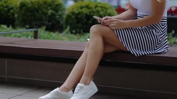 Joven chica hermosa con una camisa blanca y zapatillas blancas sentadas en la acera, sosteniendo un teléfono y marcando el mensaje. Ella sonríe y es feliz . — Vídeo de stock