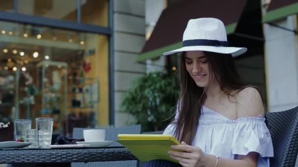 Junge attraktive Frau mit Hut, sitzt in einem Straßencafé und tippt auf Tablet. — Stockvideo
