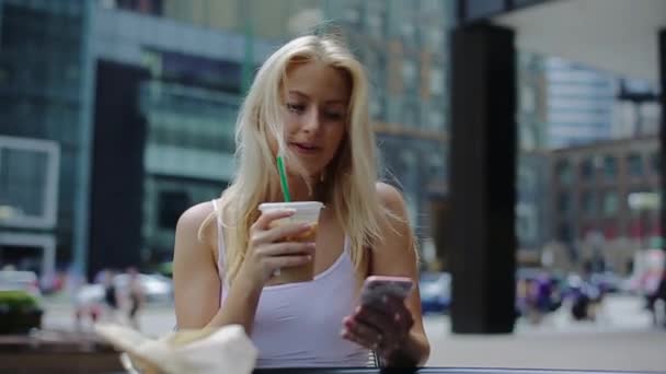 Una joven rubia sentada a la mesa, bebiendo una copa y recibiendo un mensaje por teléfono . — Vídeo de stock