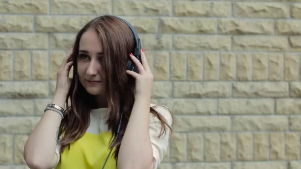 En ung flicka stående av en tegel vägg och lyssna på musik med hörlurar, Dans, glad och flirta. — Stockvideo