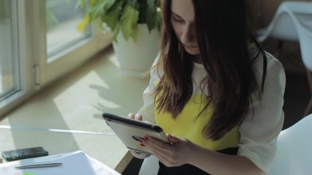 Une jeune fille est assise à une table près de la fenêtre et travaille sur la tablette. Pense, sourit, prend un stylo et prend des notes sur le papier . — Video