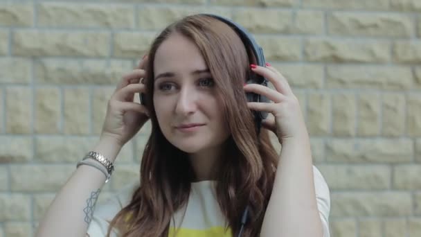 Una chica joven de pie junto a una pared de ladrillo y con auriculares. Escuchar Música, Bailar, Sonreír y Coquetear . — Vídeo de stock