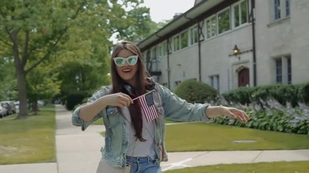 Mujer joven con gafas ondeando estrellas y rayas en frente de una casa de dos pisos en el vecindario . — Vídeos de Stock