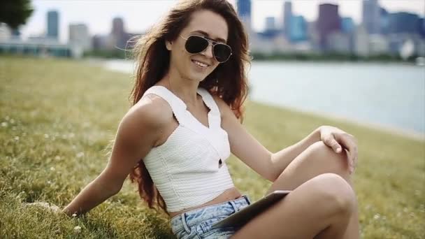 Una chica atractiva con el pelo largo morena usando una camisa blanca, pantalones cortos de Jean, y gafas de sol negras está sentado junto a un lago con una tableta — Vídeo de stock