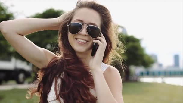 Attraktives Mädchen mit langen braunen Haaren, weißem Hemd, Jeanshose und schwarzer Sonnenbrille geht lächelnd am See entlang, während sie telefoniert — Stockvideo
