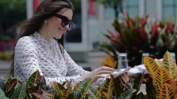 Młoda piękna kobieta w okulary siedzi w Sidewalk Cafe wśród roślin ozdobnych. — Wideo stockowe
