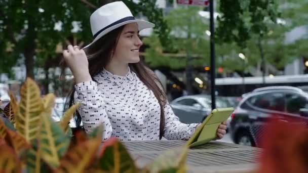 Junge schöne lächelnde Frau sitzt in einem Straßencafé in der Innenstadt, mit Tablet. — Stockvideo