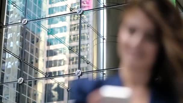 Ein junges Mädchen steht vor dem Gebäude und auf dem Handy. Hand hält Haare und lächelt. — Stockvideo