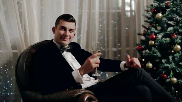 Junge brünette Kerl im Anzug sitzt in einem Stuhl neben dem Weihnachtsbaum und trinkt Champagner. — Stockvideo