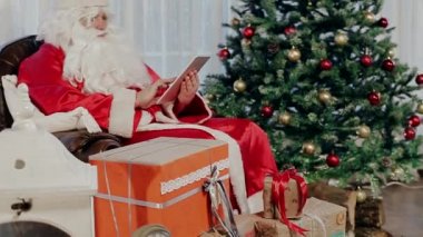 Noel Baba Noel ağacı ve bir Tablet Holding yanındaki bir sandalyede oturur.