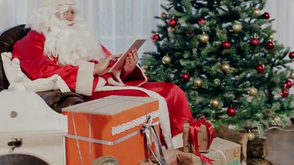 Santa Claus sedí v křesle vedle vánoční strom a drží Tablet. — Stock video