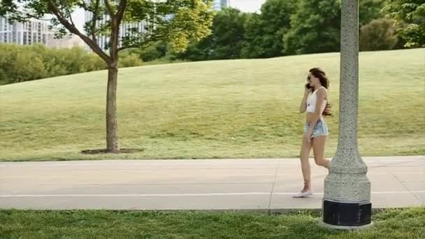 Pretty Girl Skipping With Her Cellphone in the Big Green Park With Tall Buildings in the Background in Slow Motion — Stock Video