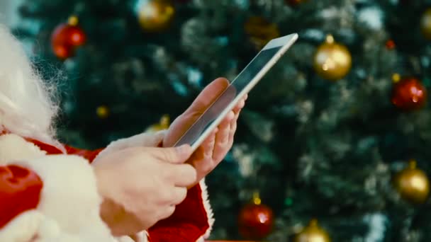 Santa Claus se nachází poblíž vánoční strom a práce na tabletu. — Stock video