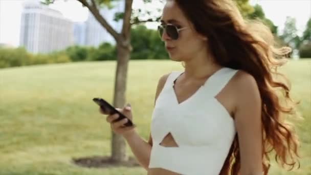 Attraktives Mädchen in weißem Hemd und Jeanshose mit schwarzer Sonnenbrille und langen lockigen Haaren, das an einem sonnigen Tag mit dem Handy in der Hand im riesigen Park tanzt — Stockvideo