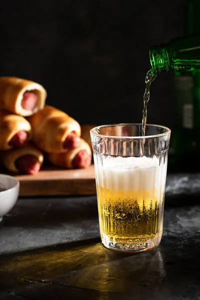 Hafif bira bardağa dökülür. Arkadaki tahtanın üzerindeki hamurda sosisler var.