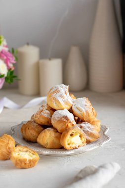 Beyaz bir masada gri bir tabakta kremalı ev yapımı profiteroller. Taze çiçekli sepet.