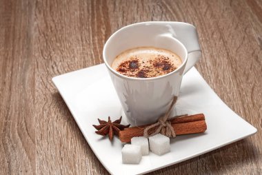 Dikdörtgen şeklinde bir fincan üzerinde beyaz güzel bir fincan. Üzerinde rendelenmiş çikolata serpiştirilmiş sütlü sıcak kahve. Yanında üç parça şeker, bir anason yıldızı ve tarçın var..