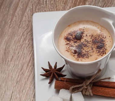 Dikdörtgen bir fincan üzerinde beyaz güzel bir fincan. Üzerine rendelenmiş çikolata serpiştirilmiş sütlü sıcak kahve. Yanında şeker, anason yıldızı ve tarçın çubukları var. Boşluğu kopyala.